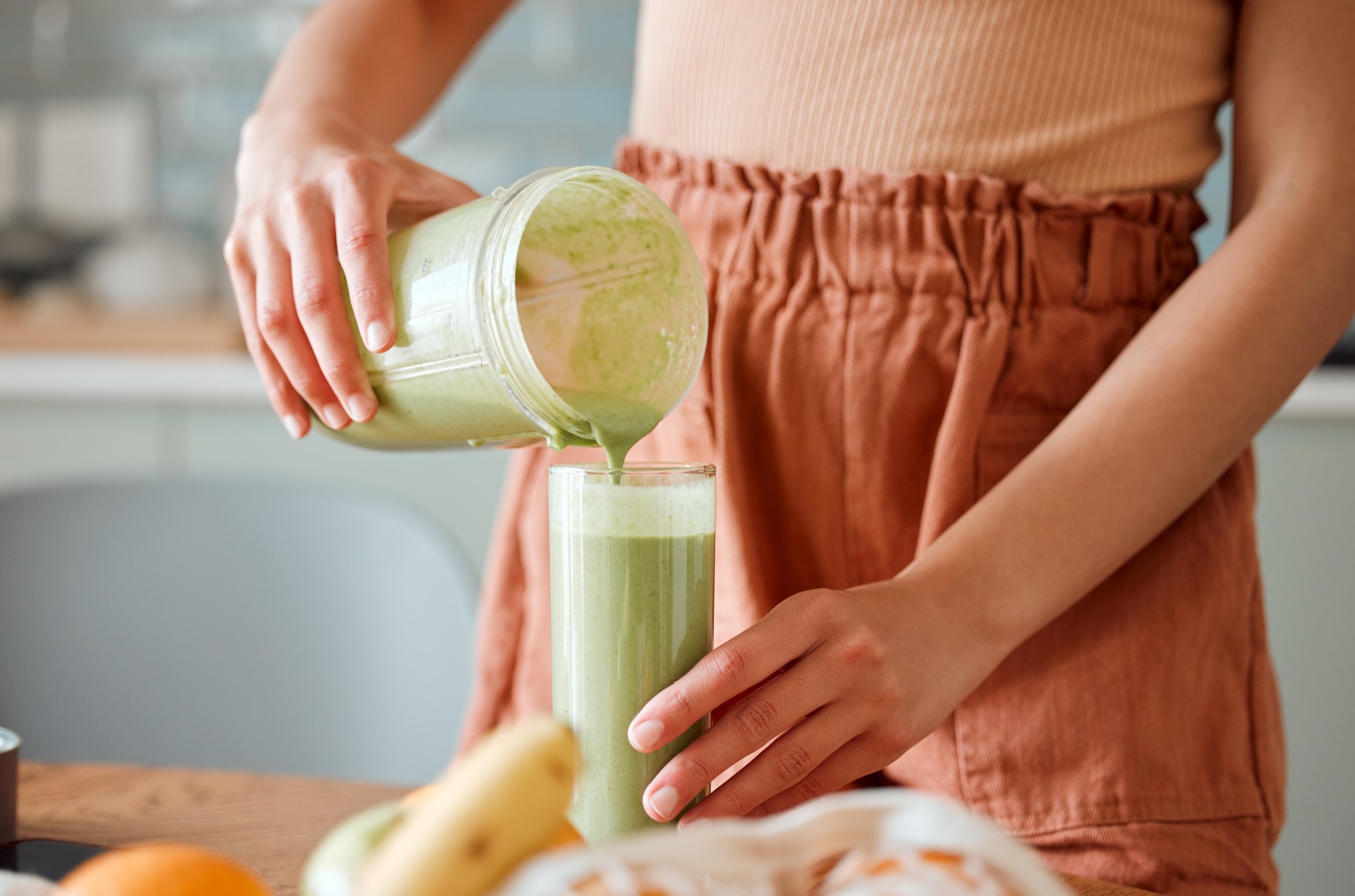 Woman pouring healthy smoothie in a glass from a blender jar on a counter for detox. Female making fresh green fruit  juice in her kitchen with vegetables and consumables for a fit lifestyle.